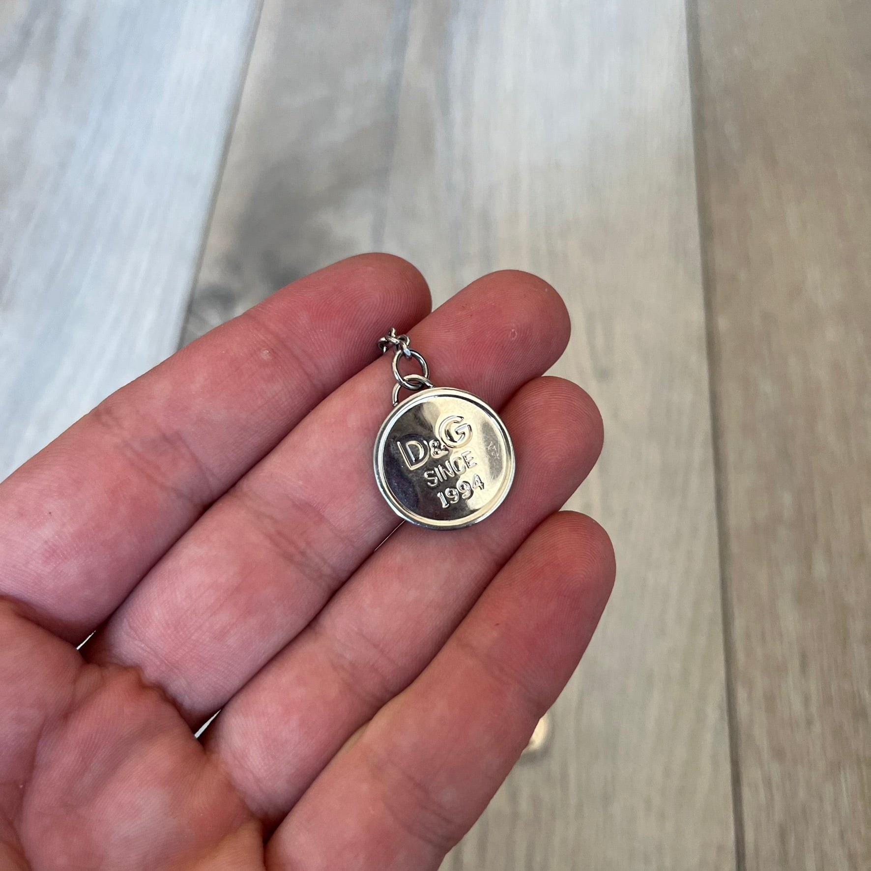 Dolce & Gabbana “D&G Since 1994” Earrings are a pair of 2000s silver-tone hanging earrings. Each earring features a short chain with a round pendant engraved with “D&G Since 1994.” The earrings have a polished metal finish and matching circular plaques.
