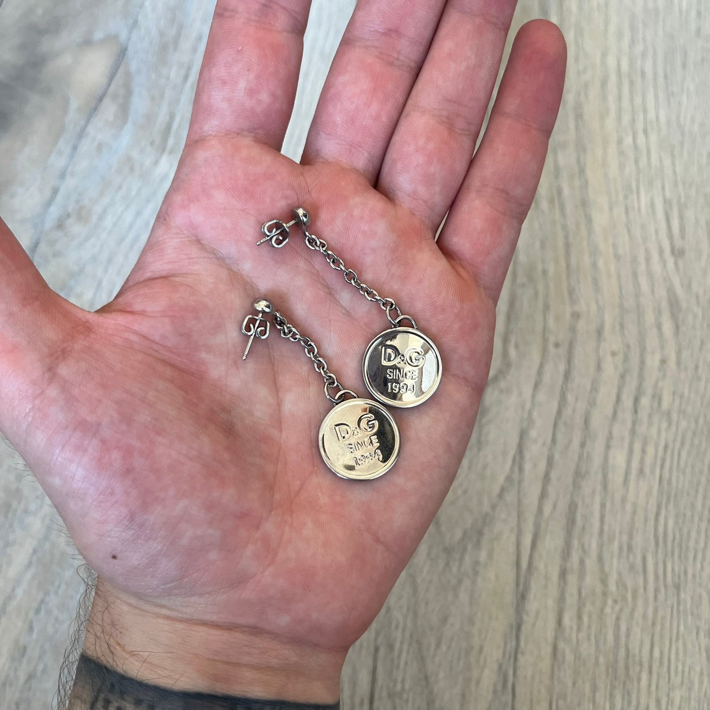 Dolce & Gabbana “D&G Since 1994” Earrings are a pair of 2000s silver-tone hanging earrings. Each earring features a short chain with a round pendant engraved with “D&G Since 1994.” The earrings have a polished metal finish and matching circular plaques.
