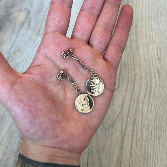 Dolce & Gabbana “D&G Since 1994” Earrings are a pair of 2000s silver-tone hanging earrings. Each earring features a short chain with a round pendant engraved with “D&G Since 1994.” The earrings have a polished metal finish and matching circular plaques.
