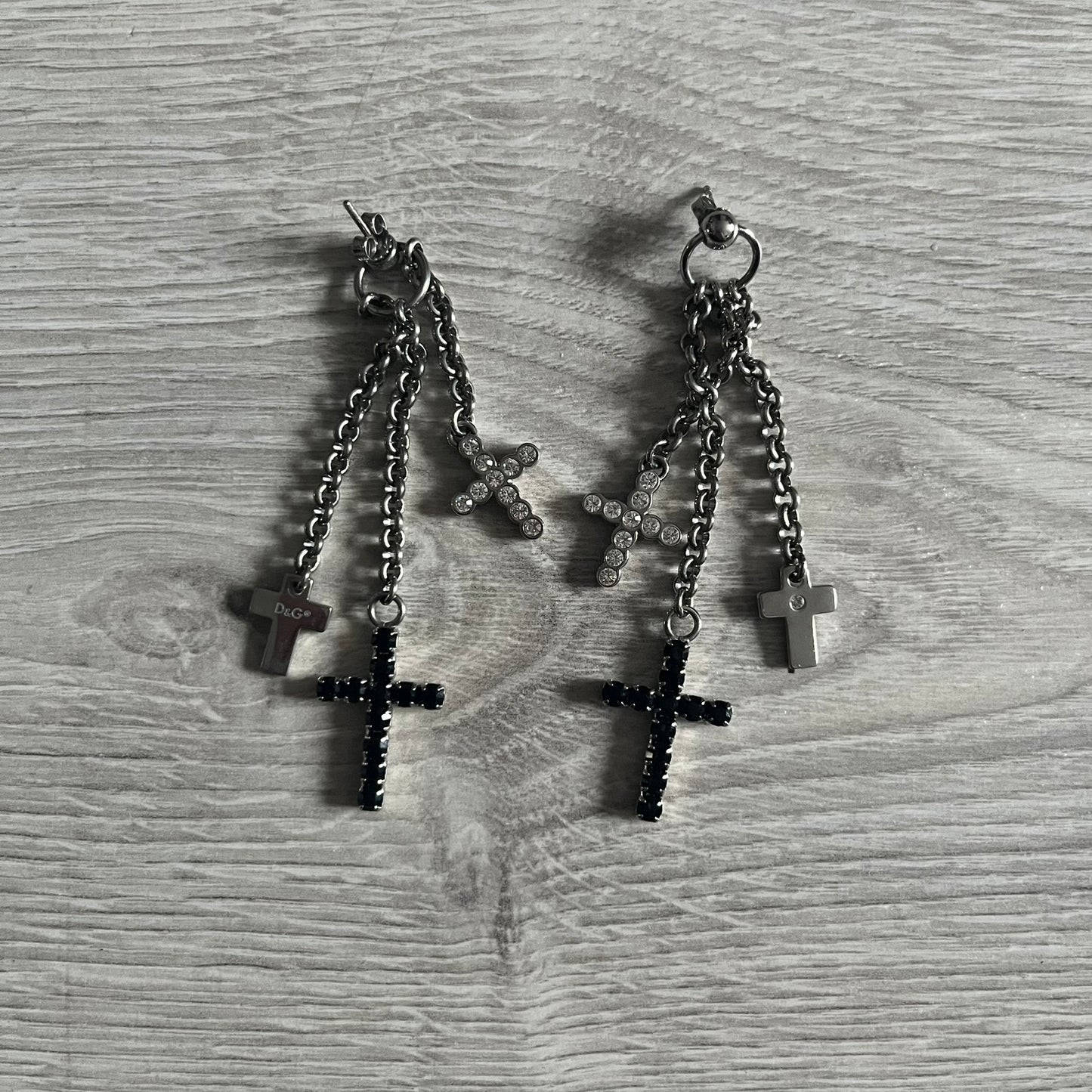 Dolce & Gabbana Dangle Drop Cross Earrings. Pair of earrings featuring three crosses of varying lengths on each, including one with black stones, one with clear stones, and one full metal cross with a D&G logo and a small clear stone. Comes with original box and dustbag