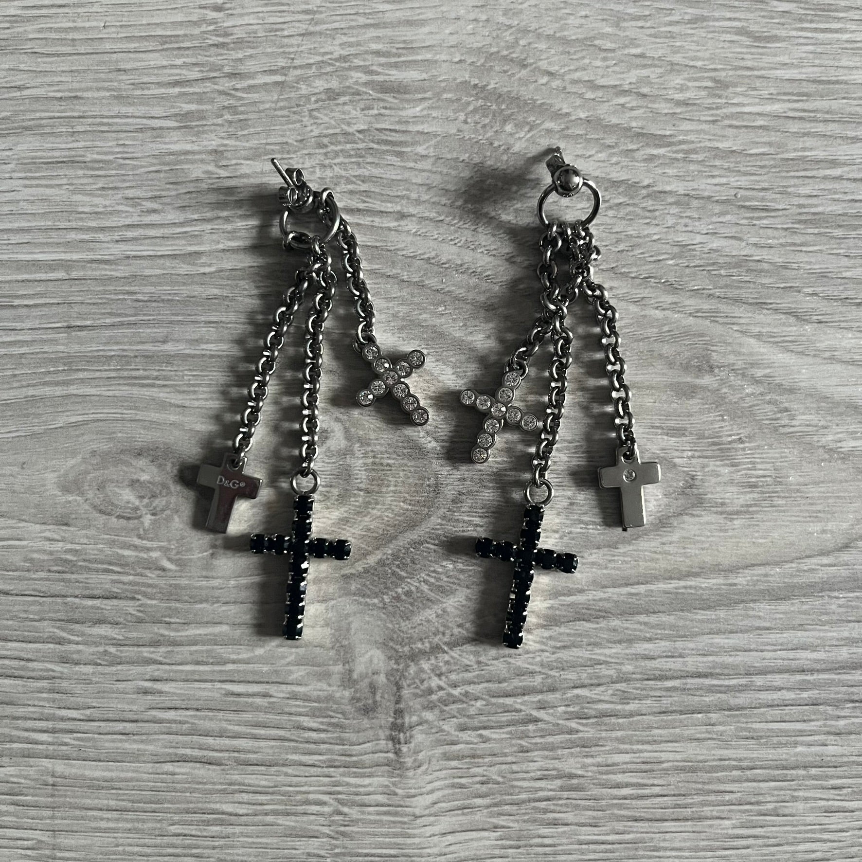 Dolce & Gabbana Dangle Drop Cross Earrings. Pair of earrings featuring three crosses of varying lengths on each, including one with black stones, one with clear stones, and one full metal cross with a D&G logo and a small clear stone. Comes with original box and dustbag