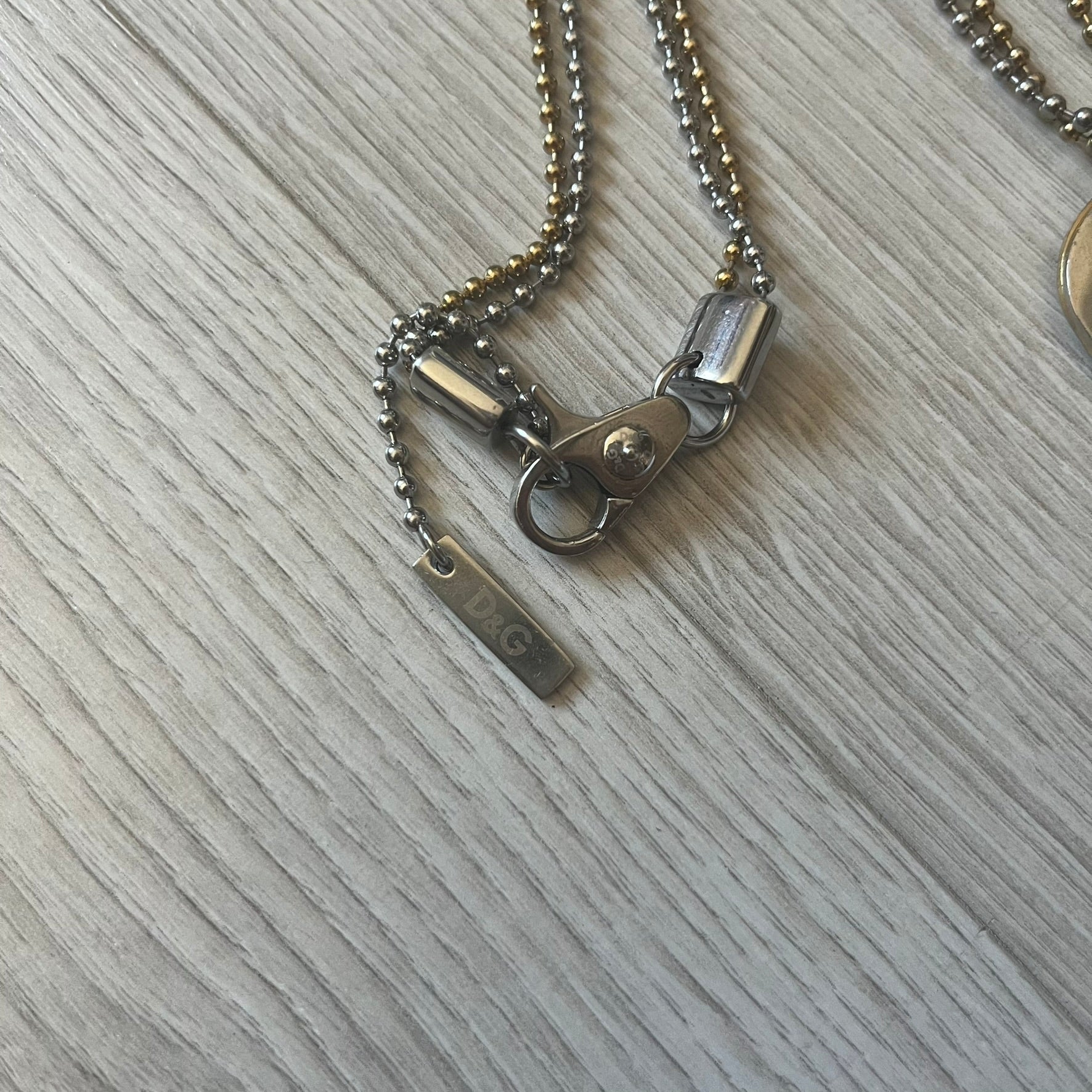 Dolce & Gabbana double dog tag necklace featuring two layered pendants: a clear gold-tone tag underneath and a silver-tone tag on top with a cut-out D&G logo revealing the gold beneath. Designed with both silver and gold chains, it creates a striking two-tone effect.
