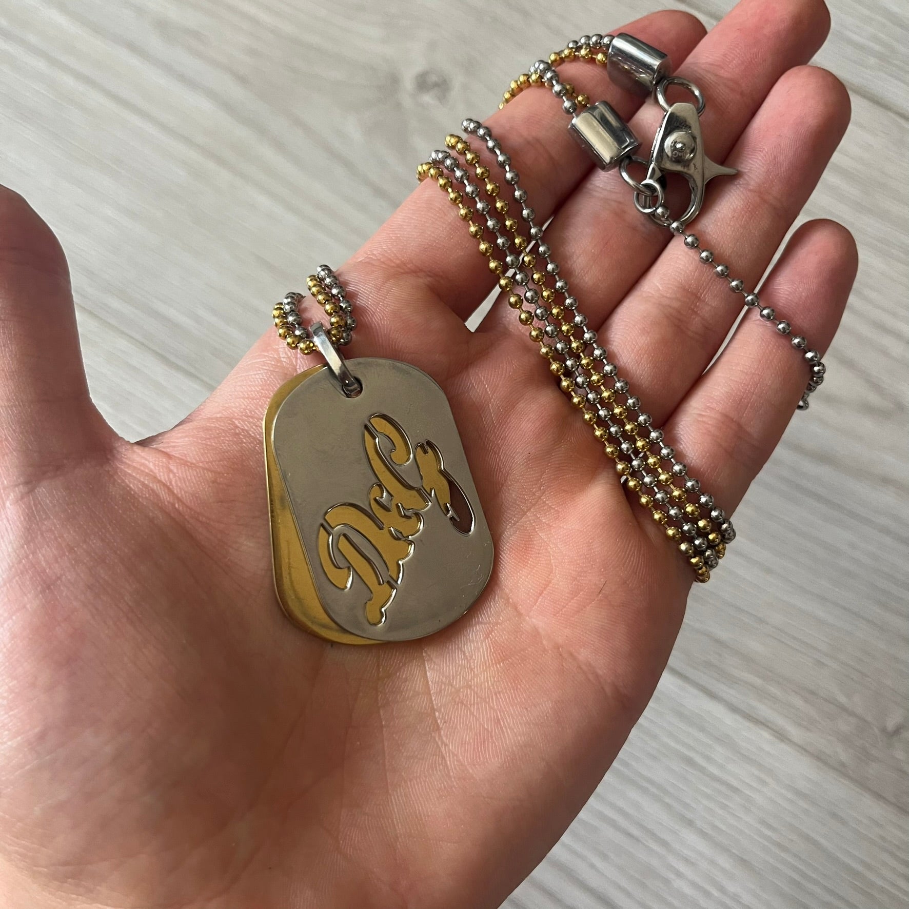 Dolce & Gabbana double dog tag necklace featuring two layered pendants: a clear gold-tone tag underneath and a silver-tone tag on top with a cut-out D&amp;G logo revealing the gold beneath. Designed with both silver and gold chains, it creates a striking two-tone effect.