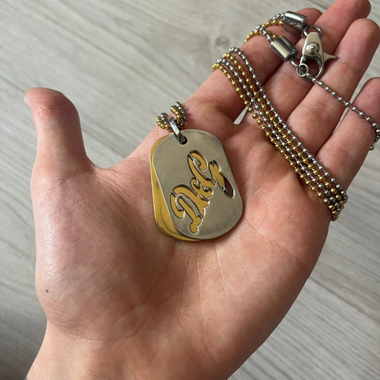Dolce &amp; Gabbana double dog tag necklace featuring two layered pendants: a clear gold-tone tag underneath and a silver-tone tag on top with a cut-out D&amp;G logo revealing the gold beneath. Designed with both silver and gold chains, it creates a striking two-tone effect.