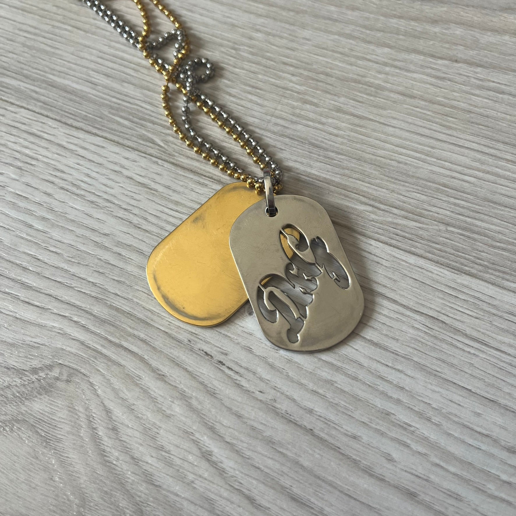 Dolce &amp; Gabbana double dog tag necklace featuring two layered pendants: a clear gold-tone tag underneath and a silver-tone tag on top with a cut-out D&amp;G logo revealing the gold beneath. Designed with both silver and gold chains, it creates a striking two-tone effect.