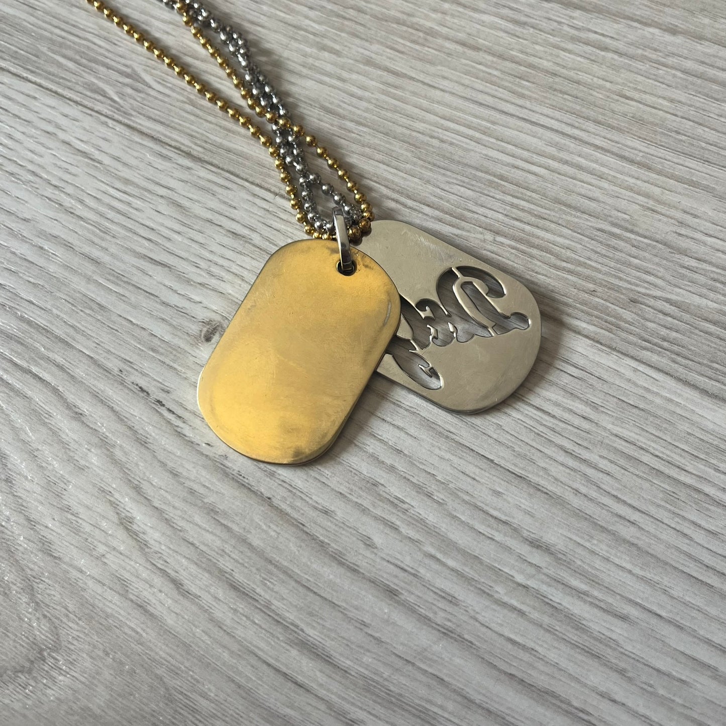 Dolce & Gabbana double dog tag necklace featuring two layered pendants: a clear gold-tone tag underneath and a silver-tone tag on top with a cut-out D&amp;G logo revealing the gold beneath. Designed with both silver and gold chains, it creates a striking two-tone effect.