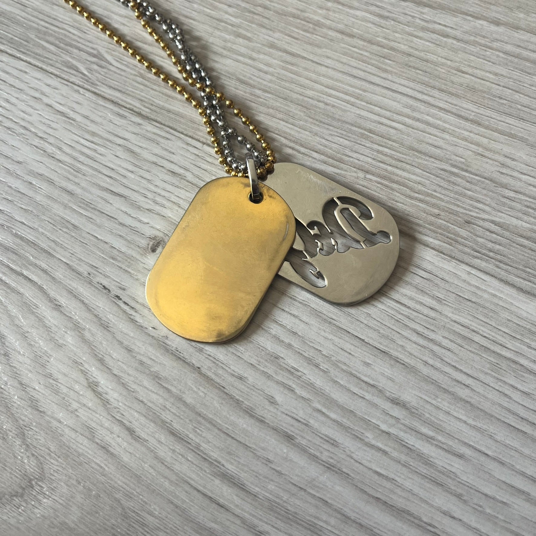 Dolce & Gabbana double dog tag necklace featuring two layered pendants: a clear gold-tone tag underneath and a silver-tone tag on top with a cut-out D&amp;G logo revealing the gold beneath. Designed with both silver and gold chains, it creates a striking two-tone effect.