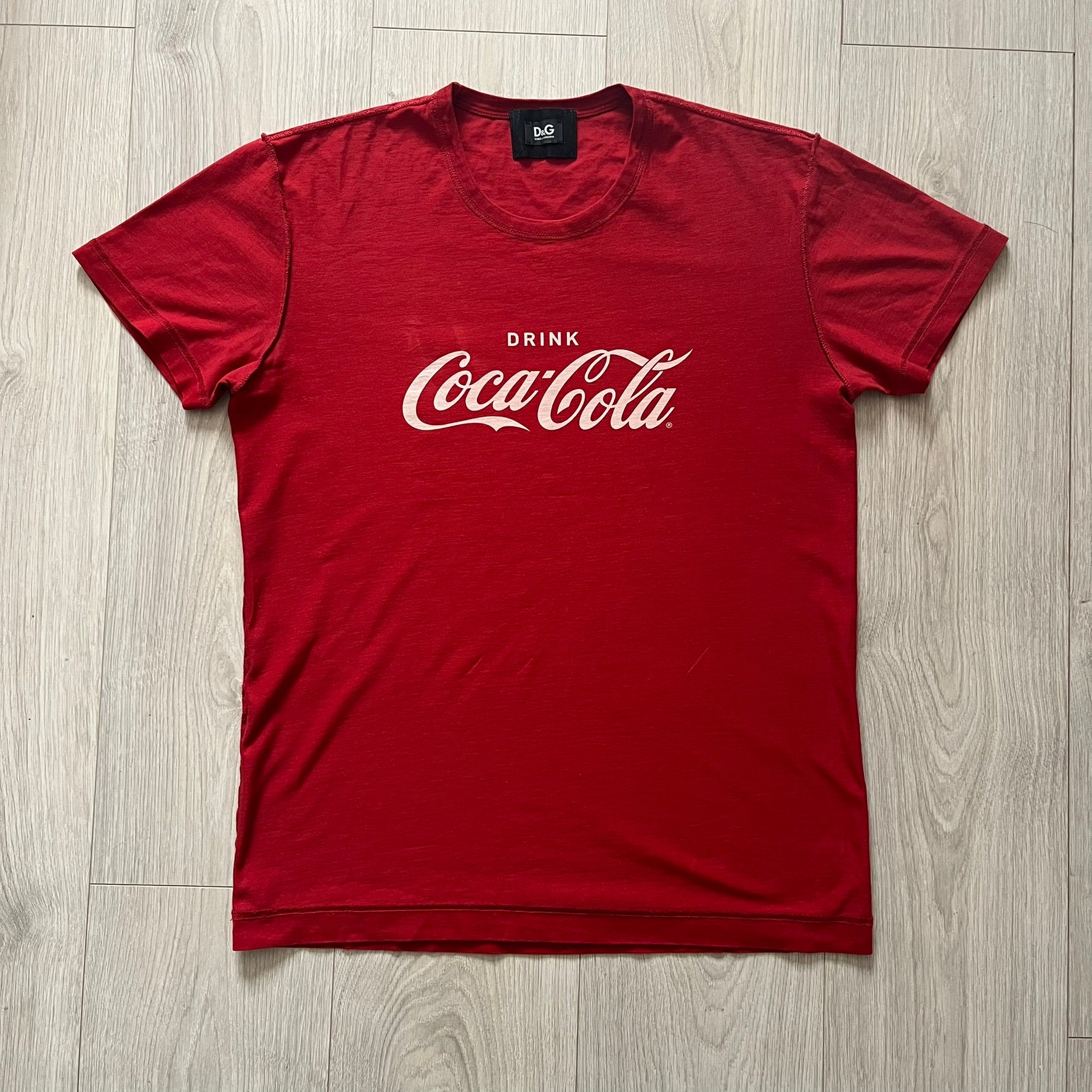 Dolce &amp; Gabbana Fall 2011 tee in vibrant red featuring a simple “Drink Coca-Cola” logo on the chest. Designed with an inside-out stitching aesthetic and a subtle back logo under the collar. A unique, collectible tee that merges casual branding with high-fashion flair.