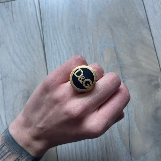 The Dolce & Gabbana Gold Logo Signet Ring is a gold-tone metal signet ring with a thick, substantial design. The top features a black background with the D&G logo in gold tone, outlined in gold. The signet is large and solid.