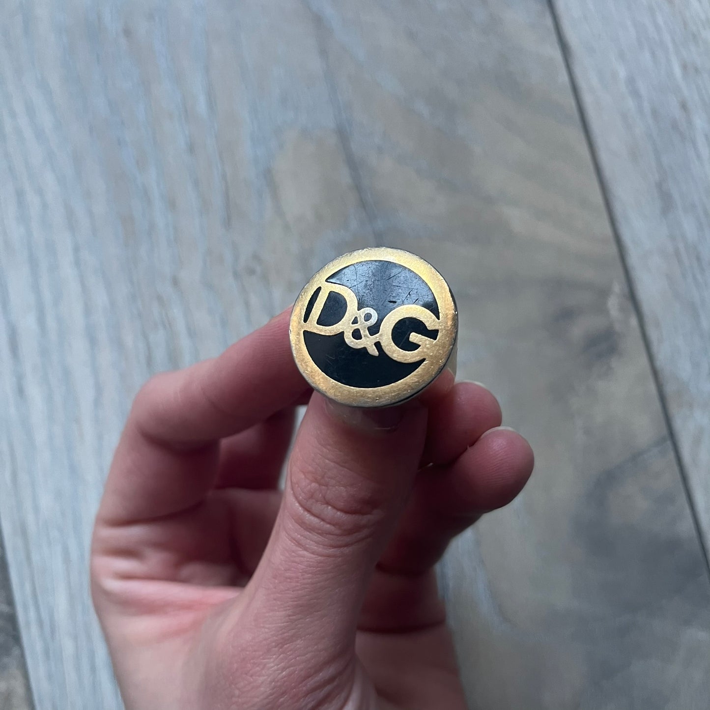 The Dolce & Gabbana Gold Logo Signet Ring is a gold-tone metal signet ring with a thick, substantial design. The top features a black background with the D&G logo in gold tone, outlined in gold. The signet is large and solid.