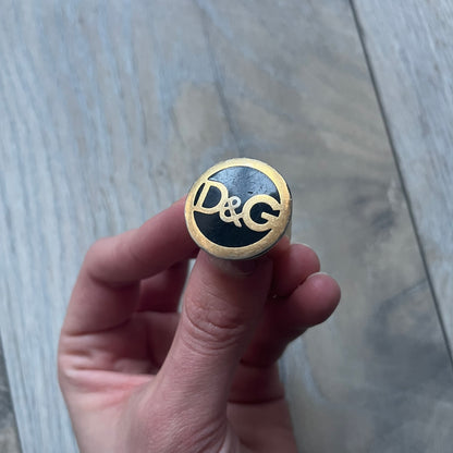 The Dolce & Gabbana Gold Logo Signet Ring is a gold-tone metal signet ring with a thick, substantial design. The top features a black background with the D&G logo in gold tone, outlined in gold. The signet is large and solid.