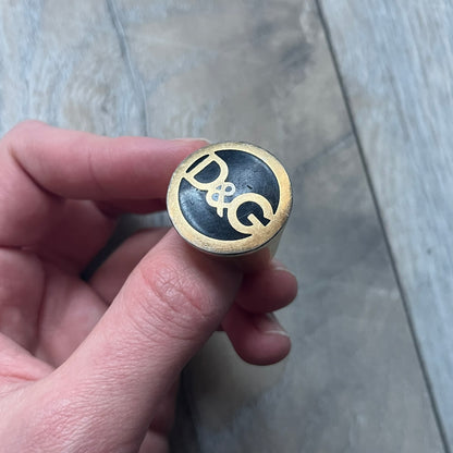 The Dolce & Gabbana Gold Logo Signet Ring is a gold-tone metal signet ring with a thick, substantial design. The top features a black background with the D&G logo in gold tone, outlined in gold. The signet is large and solid.