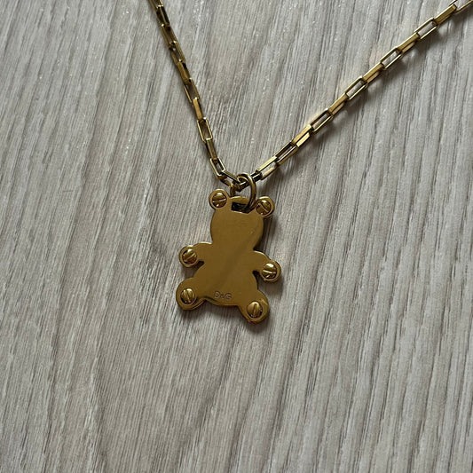 The Dolce & Gabbana Teddy Bear Necklace from the 2000s features a flat gold-tone teddy bear pendant detailed with six visible screws and a small D&G logo at the bottom. It hangs from a paperclip chain with a lobster clasp closure, it comes with a silver D&G Jewels bag.