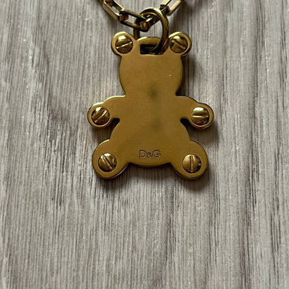 The Dolce & Gabbana Teddy Bear Necklace from the 2000s features a flat gold-tone teddy bear pendant detailed with six visible screws and a small D&G logo at the bottom. It hangs from a paperclip chain with a lobster clasp closure, it comes with a silver D&G Jewels bag.