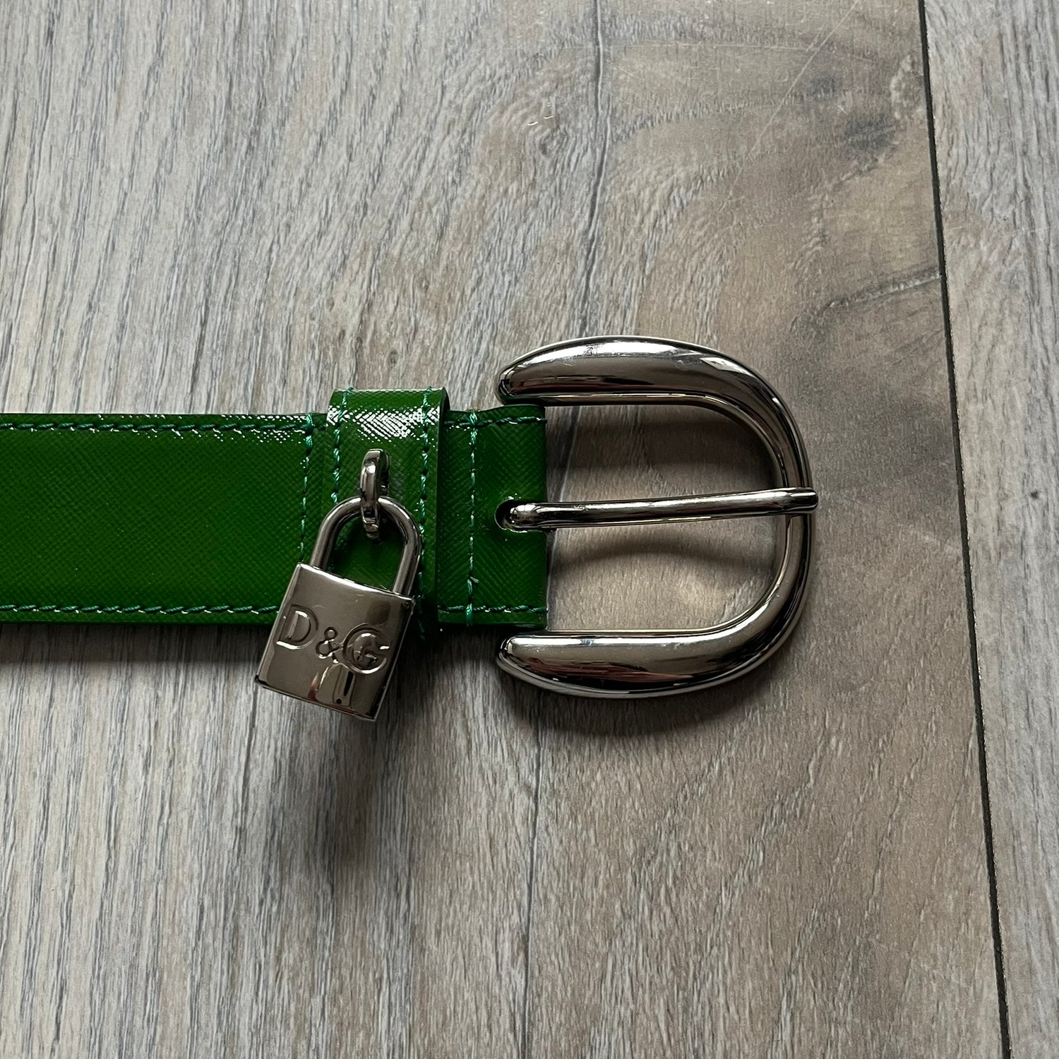 Dolce & Gabbana Green Leather Padlock Buckle Belt features a shiny green leather strap with a beige leather underside. The belt has metal-outlined eyelets for the holes and a clean, simple silver-tone buckle. Next to the buckle is a silver-tone padlock engraved with D&G, along with a fixed metal loop at the center of the belt intended to attach a chain (chain not included).
