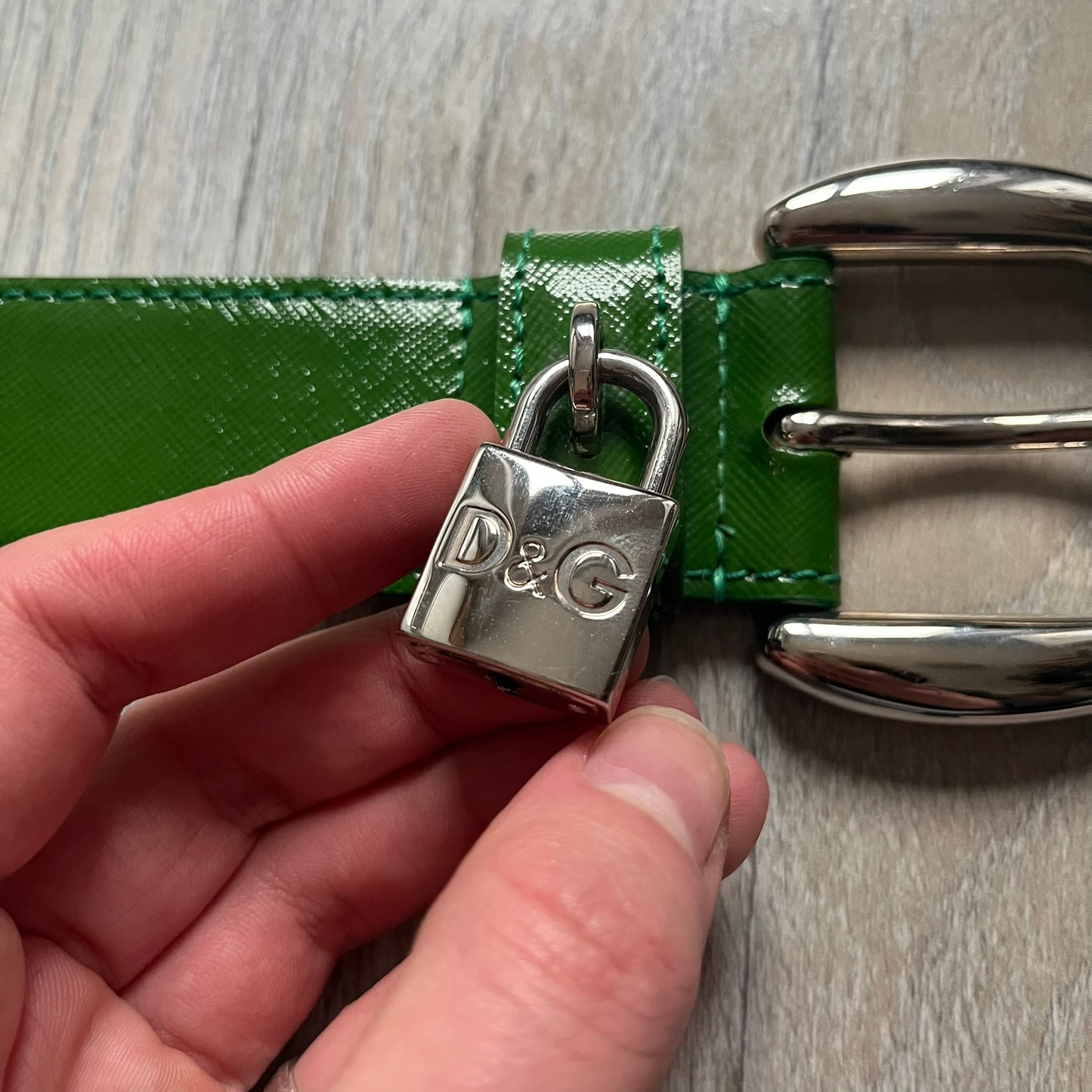 Dolce & Gabbana Green Leather Padlock Buckle Belt features a shiny green leather strap with a beige leather underside. The belt has metal-outlined eyelets for the holes and a clean, simple silver-tone buckle. Next to the buckle is a silver-tone padlock engraved with D&G, along with a fixed metal loop at the center of the belt intended to attach a chain (chain not included).