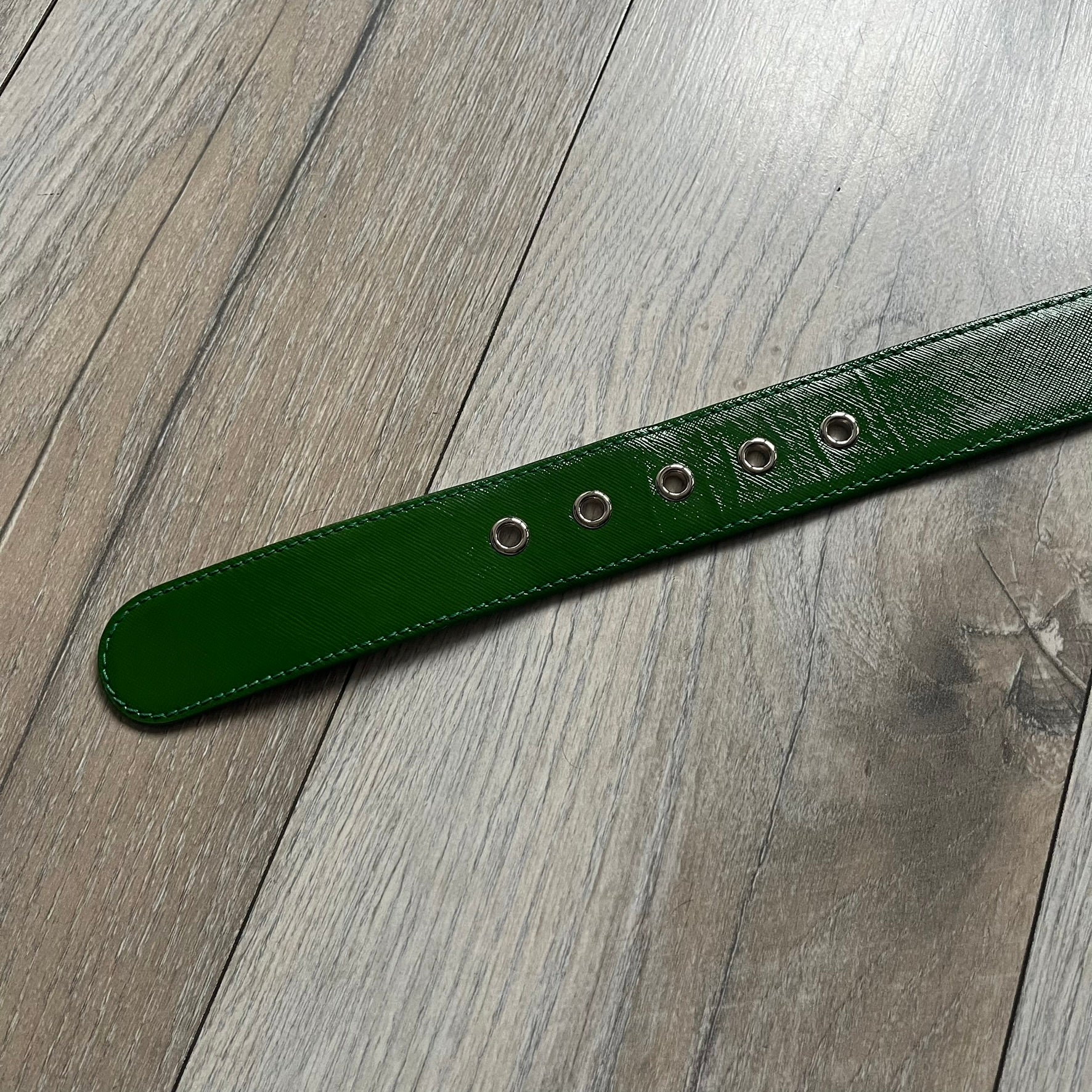 Dolce & Gabbana Green Leather Padlock Buckle Belt features a shiny green leather strap with a beige leather underside. The belt has metal-outlined eyelets for the holes and a clean, simple silver-tone buckle. Next to the buckle is a silver-tone padlock engraved with D&G, along with a fixed metal loop at the center of the belt intended to attach a chain (chain not included).