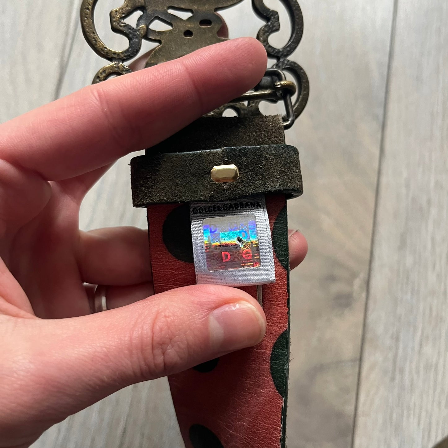 Dolce & Gabbana Green Suede Hearts Belt is made from very dark green suede with an orange leather interior featuring large dot detailing. The buckle is a metal D&G logo surrounded by eight heart shapes pointing outward, all finished in a dark, aged-looking metal tone. The belt comes with the original hologram tag attached.