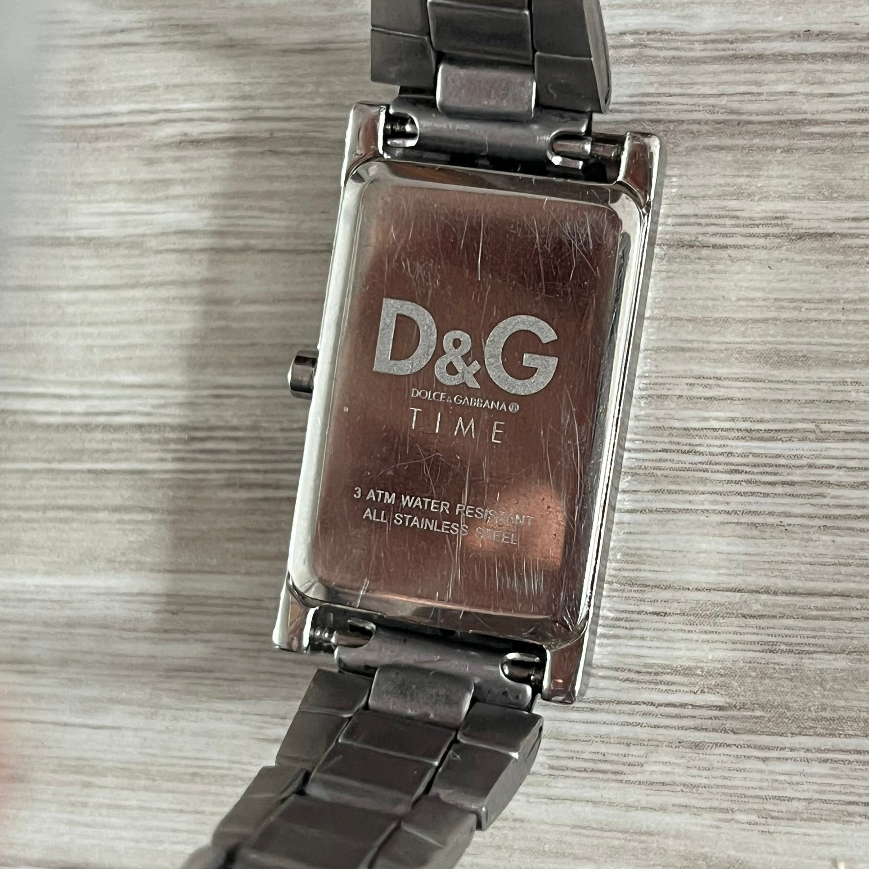  Dolce &amp; Gabbana Holographic Face Analog Watch featuring a slim stainless steel case and a dark face with repeating brown holographic D&amp;G logos. The minimalist dial includes just the small D&amp;G logo and two hands for hours and minutes, giving it a clean and refined look. Water resistant up to 3 ATM, making it both stylish and functional for everyday wear.