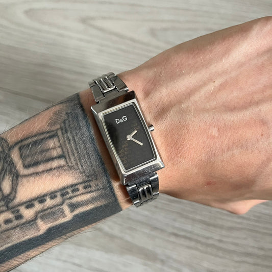 Dolce &amp; Gabbana Holographic Face Analog Watch featuring a slim stainless steel case and a dark face with repeating brown holographic D&amp;G logos. The minimalist dial includes just the small D&amp;G logo and two hands for hours and minutes, giving it a clean and refined look. Water resistant up to 3 ATM, making it both stylish and functional for everyday wear.