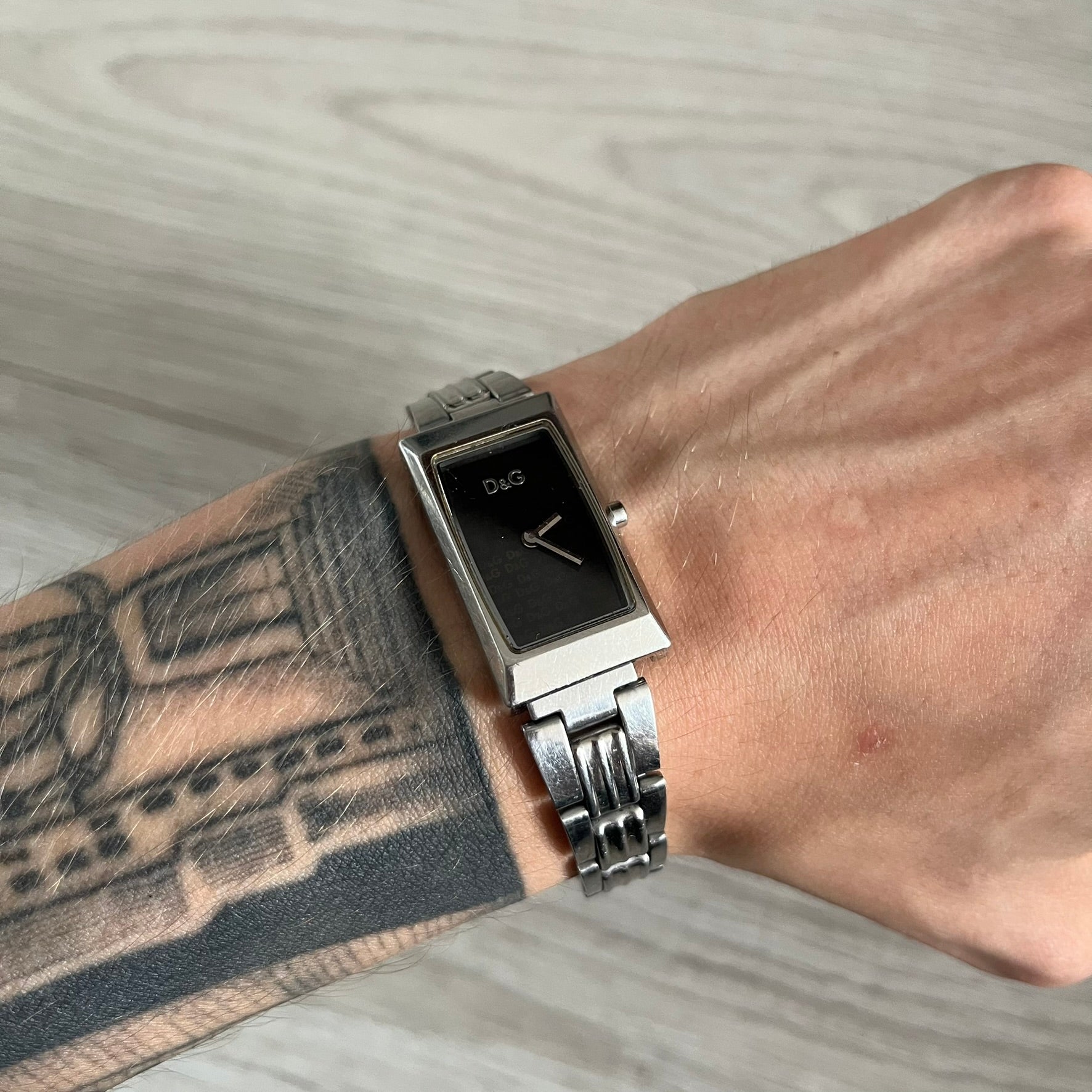 Dolce &amp; Gabbana Holographic Face Analog Watch featuring a slim stainless steel case and a dark face with repeating brown holographic D&amp;G logos. The minimalist dial includes just the small D&amp;G logo and two hands for hours and minutes, giving it a clean and refined look. Water resistant up to 3 ATM, making it both stylish and functional for everyday wear.