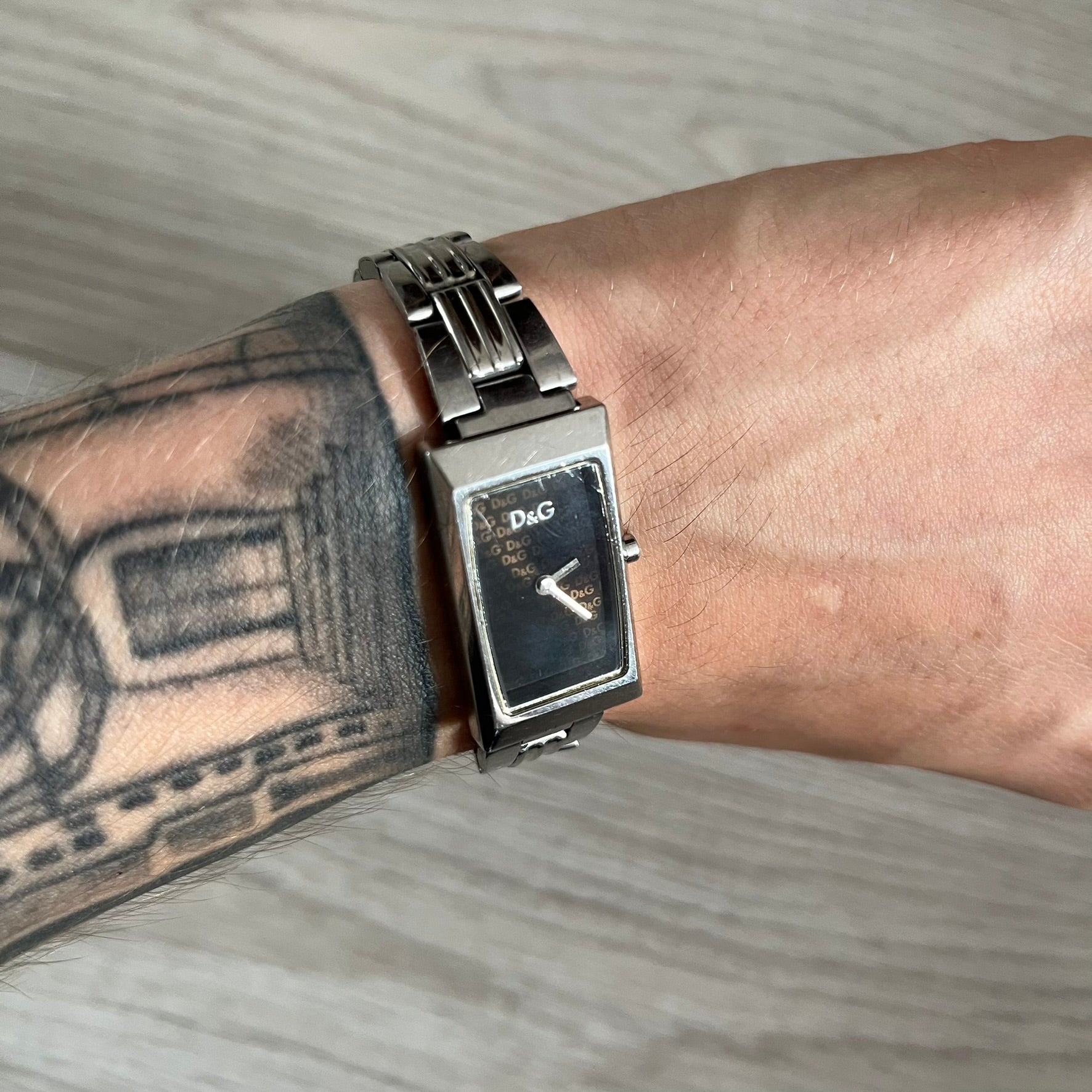  Dolce &amp; Gabbana Holographic Face Analog Watch featuring a slim stainless steel case and a dark face with repeating brown holographic D&amp;G logos. The minimalist dial includes just the small D&amp;G logo and two hands for hours and minutes, giving it a clean and refined look. Water resistant up to 3 ATM, making it both stylish and functional for everyday wear.