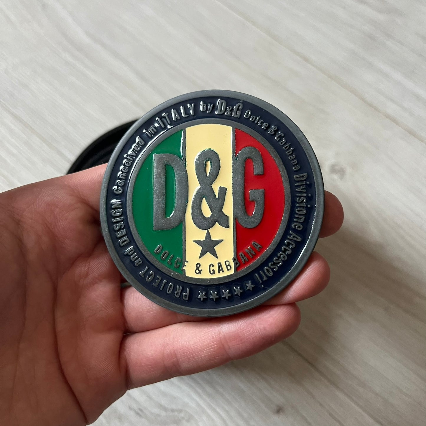 2000's Dolce & Gabbana black leather belt featuring a circular dark silver-tone metal buckle with the Italian flag and D&G logo in the center. The buckle is bordered by a navy ring with engraved text reading “Project And Design Conceived In Italy By Dolce & Gabbana”.