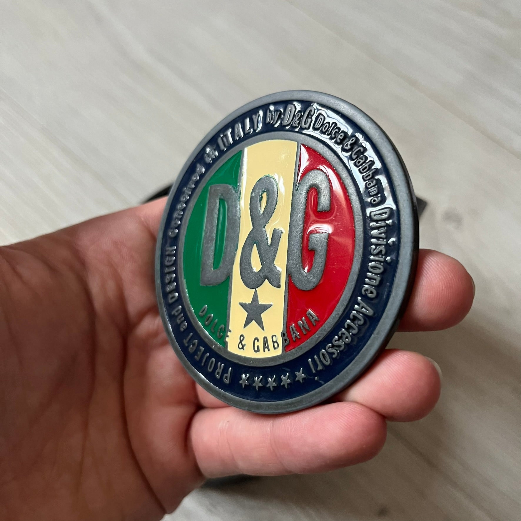 2000's Dolce & Gabbana black leather belt featuring a circular dark silver-tone metal buckle with the Italian flag and D&G logo in the center. The buckle is bordered by a navy ring with engraved text reading “Project And Design Conceived In Italy By Dolce & Gabbana”.