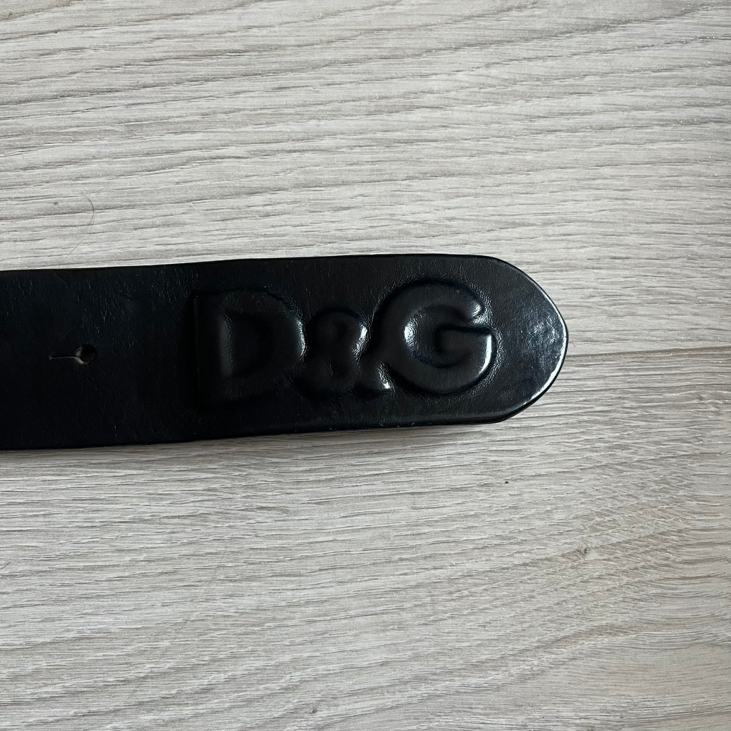 2000's Dolce & Gabbana black leather belt featuring a circular dark silver-tone metal buckle with the Italian flag and D&G logo in the center. The buckle is bordered by a navy ring with engraved text reading “Project And Design Conceived In Italy By Dolce & Gabbana”.