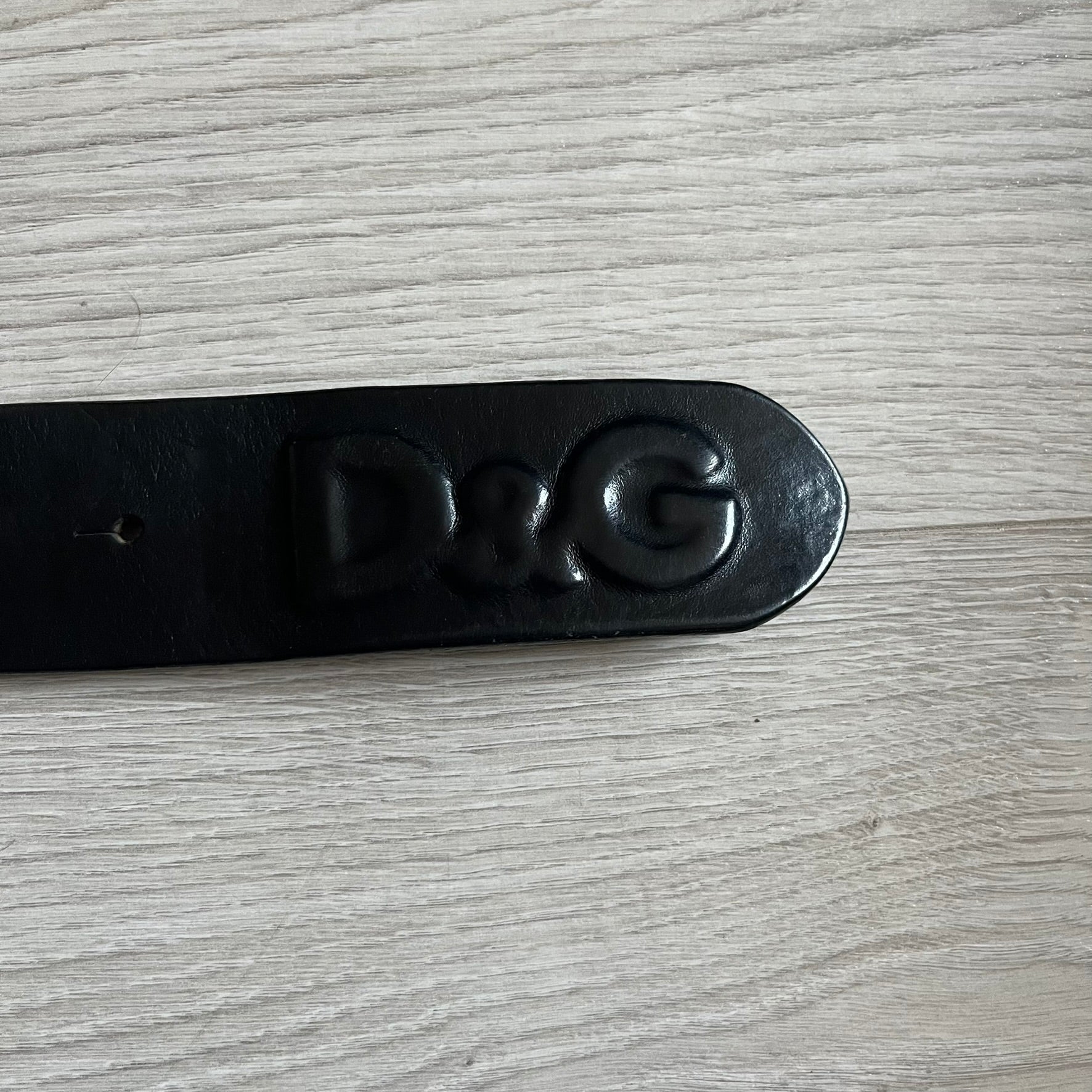 2000's Dolce & Gabbana black leather belt featuring a circular dark silver-tone metal buckle with the Italian flag and D&G logo in the center. The buckle is bordered by a navy ring with engraved text reading “Project And Design Conceived In Italy By Dolce & Gabbana”.