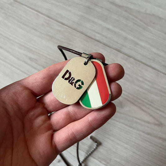 Dolce &amp; Gabbana double dog tag necklace featuring two layered pendants: a italy flag tag underneath and a silver-tone tag on top with a cut-out D&amp;G logo revealing the tricolor tag beneath. Designed with brown leather-ish string.