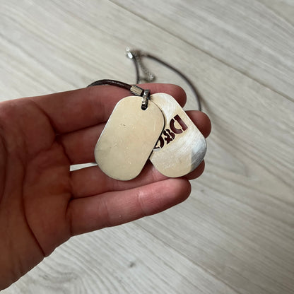 Dolce &amp; Gabbana double dog tag necklace featuring two layered pendants: a italy flag tag underneath and a silver-tone tag on top with a cut-out D&amp;G logo revealing the tricolor tag beneath. Designed with brown leather-ish string.
