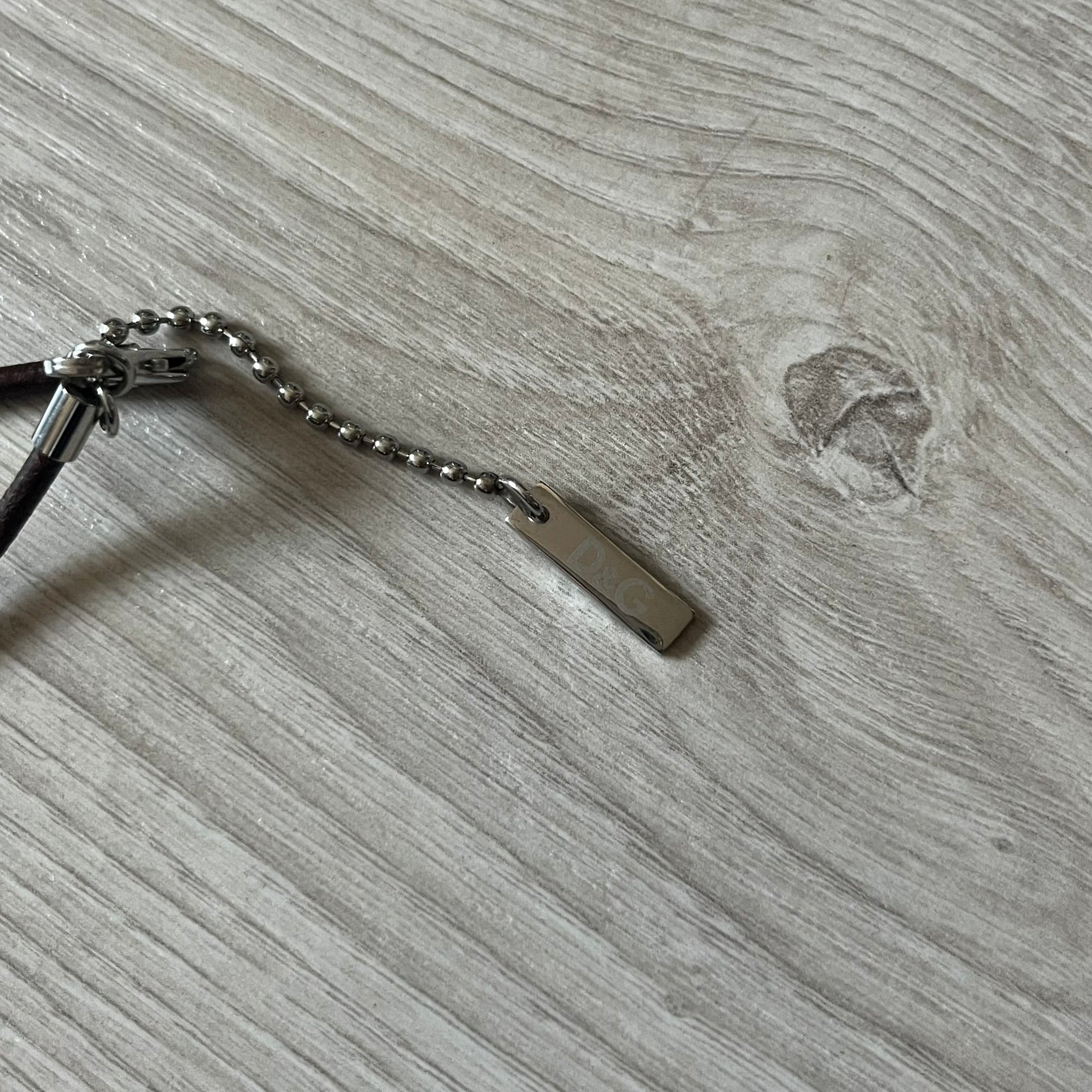 Dolce &amp; Gabbana double dog tag necklace featuring two layered pendants: a italy flag tag underneath and a silver-tone tag on top with a cut-out D&amp;G logo revealing the tricolor tag beneath. Designed with brown leather-ish string.