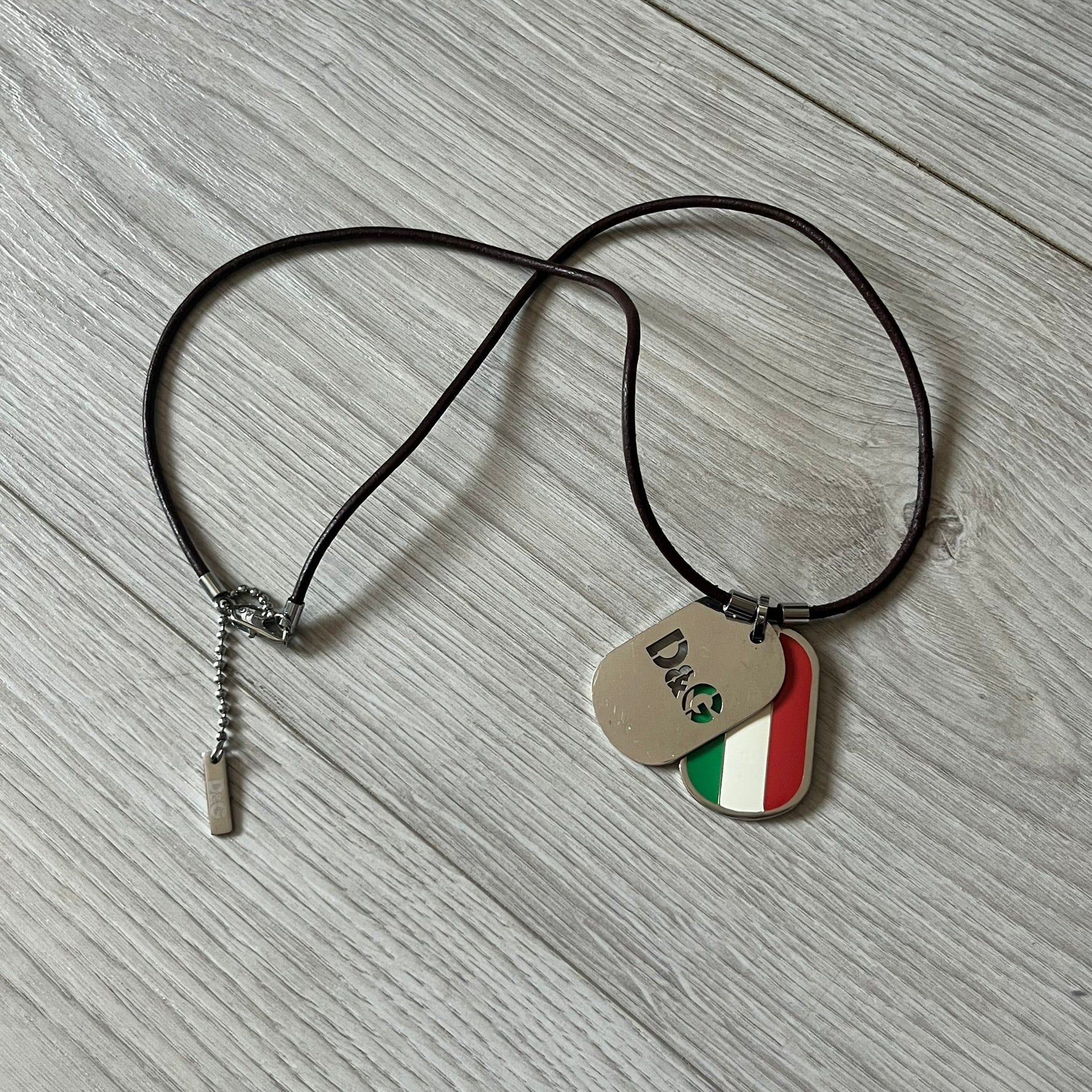 Dolce &amp; Gabbana double dog tag necklace featuring two layered pendants: a italy flag tag underneath and a silver-tone tag on top with a cut-out D&amp;G logo revealing the tricolor tag beneath. Designed with brown leather-ish string.