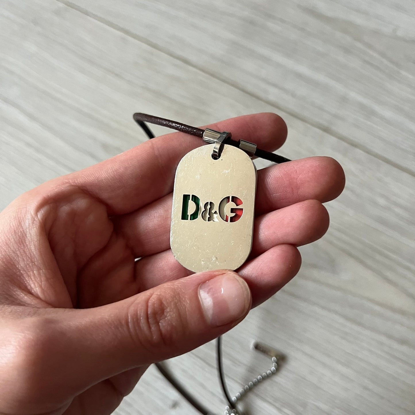 Dolce &amp; Gabbana double dog tag necklace featuring two layered pendants: a italy flag tag underneath and a silver-tone tag on top with a cut-out D&amp;G logo revealing the tricolor tag beneath. Designed with brown leather-ish string.