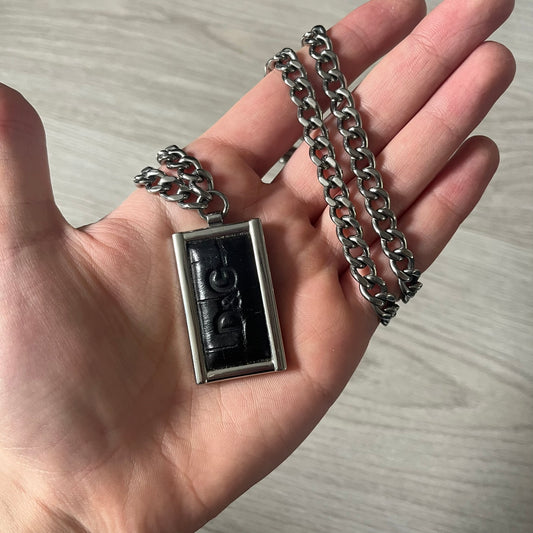 D&amp;G DJ0564 Pendant Necklace showcases a large silver curb chain paired with a rectangular silver frame pendant. Inside, black leather embossed with the D&amp;G logo adds contrast and sophistication. A striking blend of luxury leather and stainless steel craftsmanship.