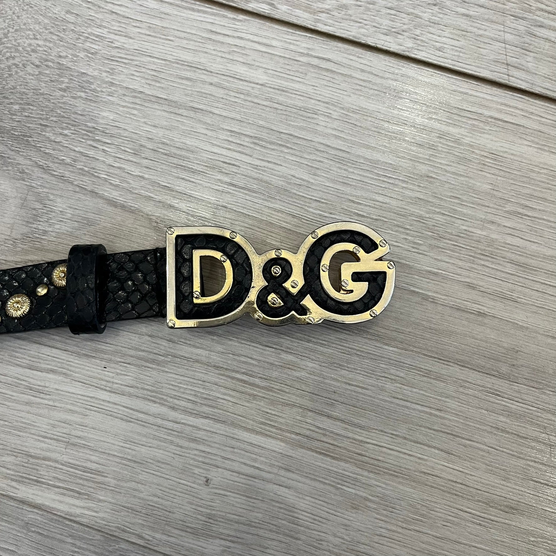 Dolce &amp; Gabbana Leather Studded Belt in black textured leather with a scaled, snake-like finish. The belt features gold stud detailing arranged in a clean pattern and a matching black D&amp;G buckle trimmed with gold hardware and exposed screws. The interior is finished in gold, adding a refined contrast to the overall design.