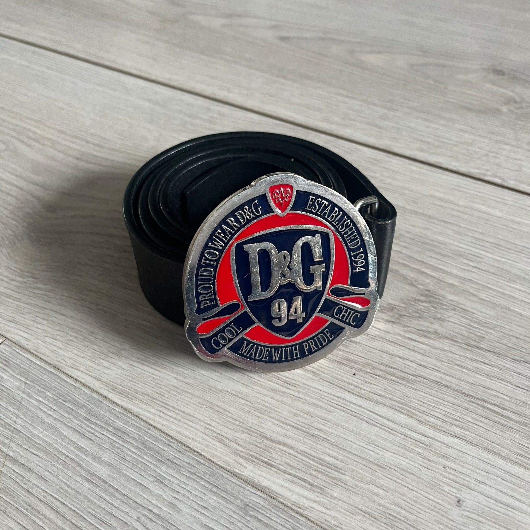 Dolce &amp; Gabbana "Made with Pride" Belt crafted from smooth black Italian leather. Features a circular silver buckle with D&amp;G 94 at the center, surrounded by “Proud to Wear D&amp;G,” “Made with Pride,” and “Established 1994” text. Finished with subtle red and navy accents, combining classic craftsmanship with contemporary flair.
