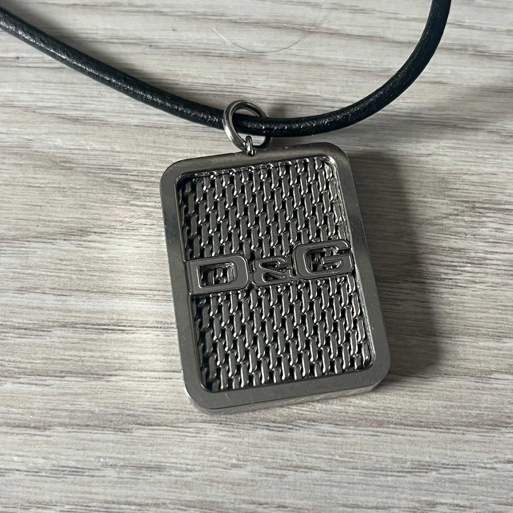 Dolce &amp; Gabbana Mesh Necklace featuring a stainless steel plate engraved with the D&amp;G logo, attached to a black leather cord. The reverse side displays a detailed grid-style mesh design. Model DJ0452, crafted with precision and industrial-inspired elegance.