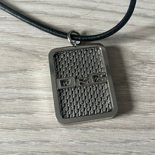 Dolce &amp; Gabbana Mesh Necklace featuring a stainless steel plate engraved with the D&amp;G logo, attached to a black leather cord. The reverse side displays a detailed grid-style mesh design. Model DJ0452, crafted with precision and industrial-inspired elegance.