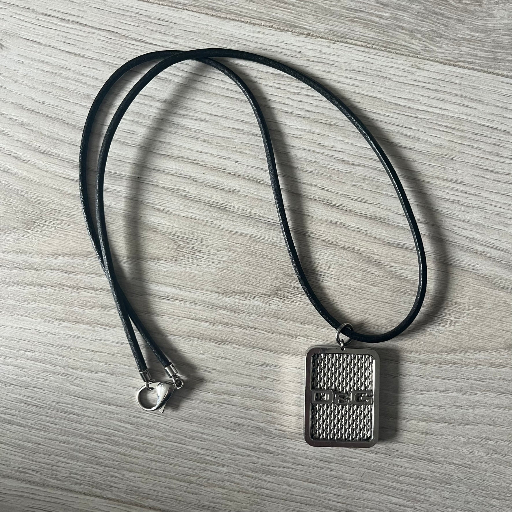 Dolce &amp; Gabbana Mesh Necklace featuring a stainless steel plate engraved with the D&amp;G logo, attached to a black leather cord. The reverse side displays a detailed grid-style mesh design. Model DJ0452, crafted with precision and industrial-inspired elegance.
