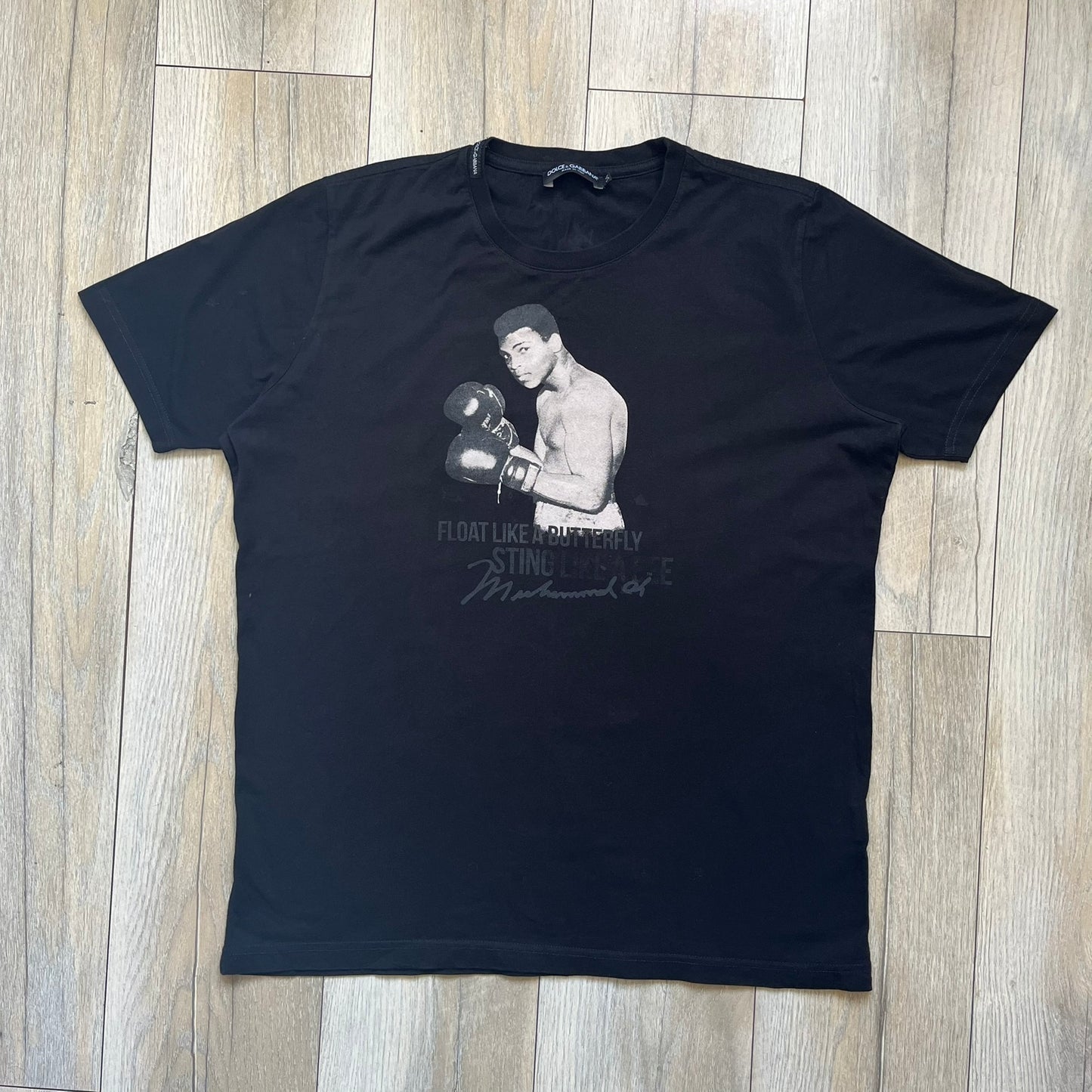  Dolce &amp; Gabbana Muhammad Ali Black Tee is a black T-shirt made in Italy from 100% cotton. The front features a grayscale portrait of Muhammad Ali. Beneath the image is the quote “float like a butterfly sting like a bee” along with a Muhammad Ali signature. 