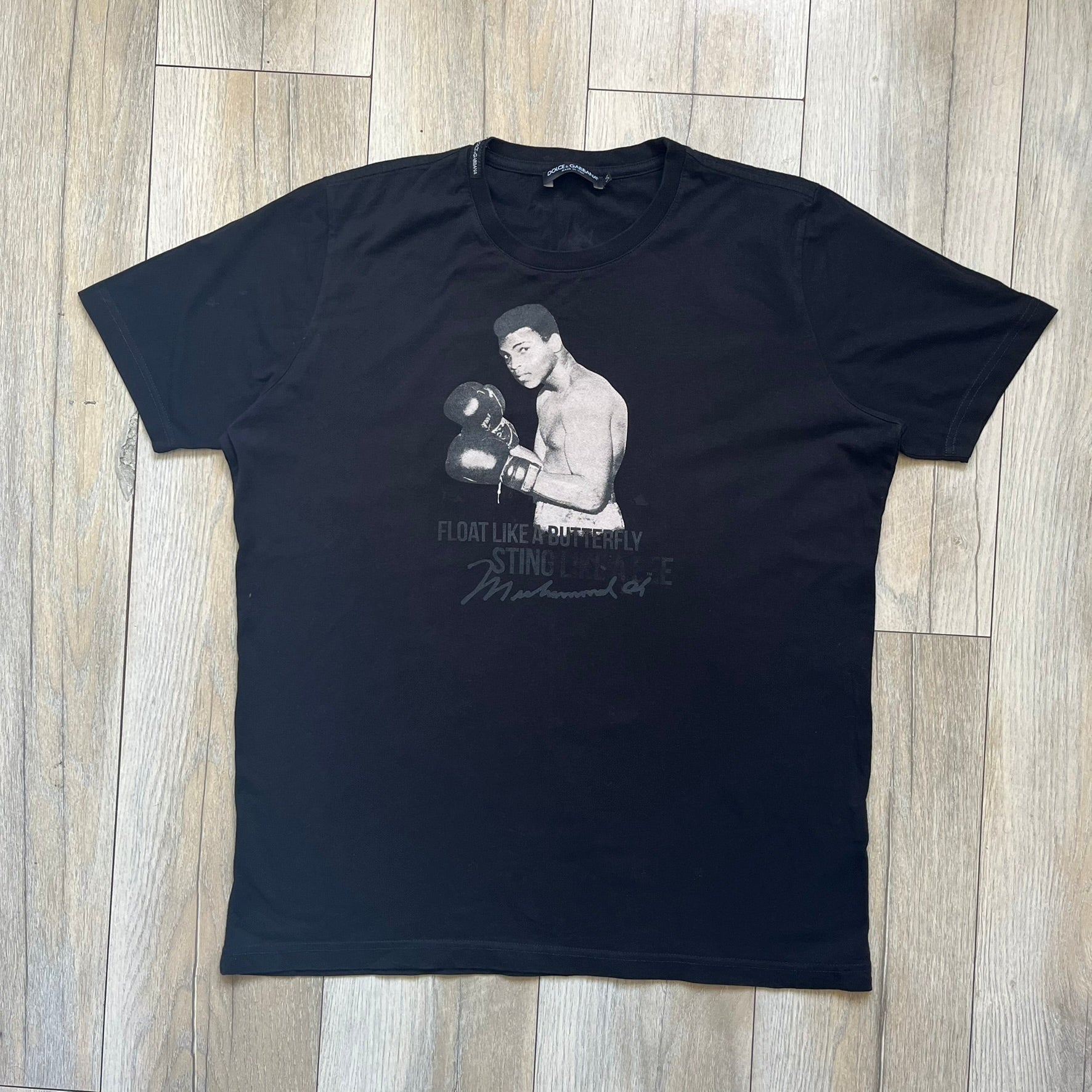  Dolce &amp; Gabbana Muhammad Ali Black Tee is a black T-shirt made in Italy from 100% cotton. The front features a grayscale portrait of Muhammad Ali. Beneath the image is the quote “float like a butterfly sting like a bee” along with a Muhammad Ali signature. 