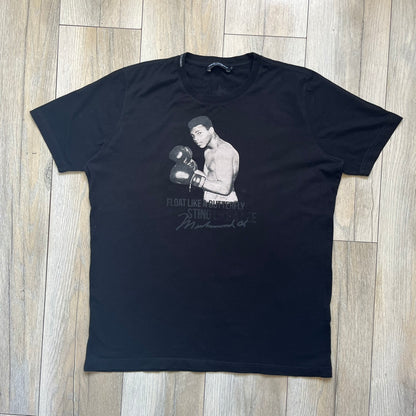  Dolce &amp; Gabbana Muhammad Ali Black Tee is a black T-shirt made in Italy from 100% cotton. The front features a grayscale portrait of Muhammad Ali. Beneath the image is the quote “float like a butterfly sting like a bee” along with a Muhammad Ali signature. 