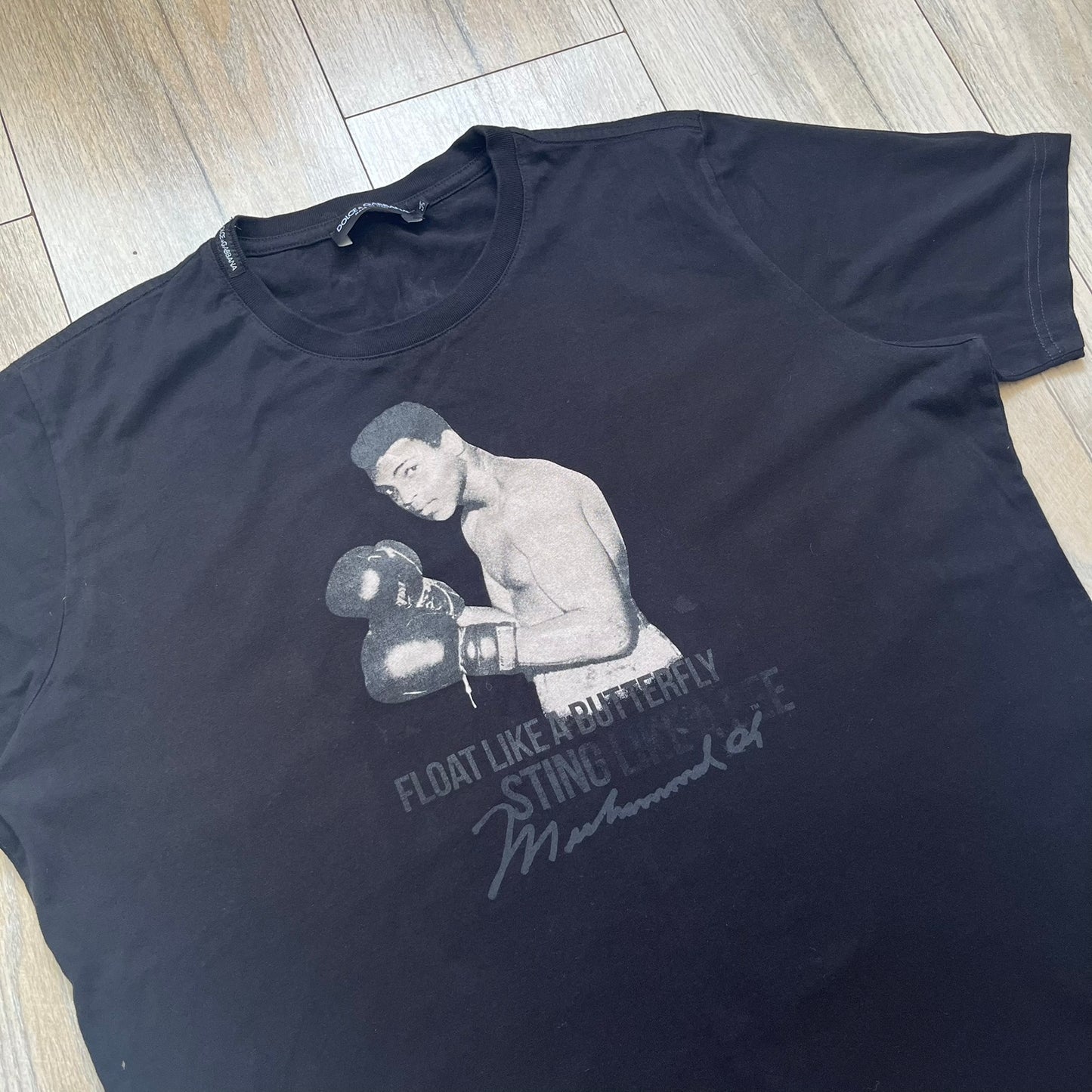  Dolce &amp; Gabbana Muhammad Ali Black Tee is a black T-shirt made in Italy from 100% cotton. The front features a grayscale portrait of Muhammad Ali. Beneath the image is the quote “float like a butterfly sting like a bee” along with a Muhammad Ali signature. 