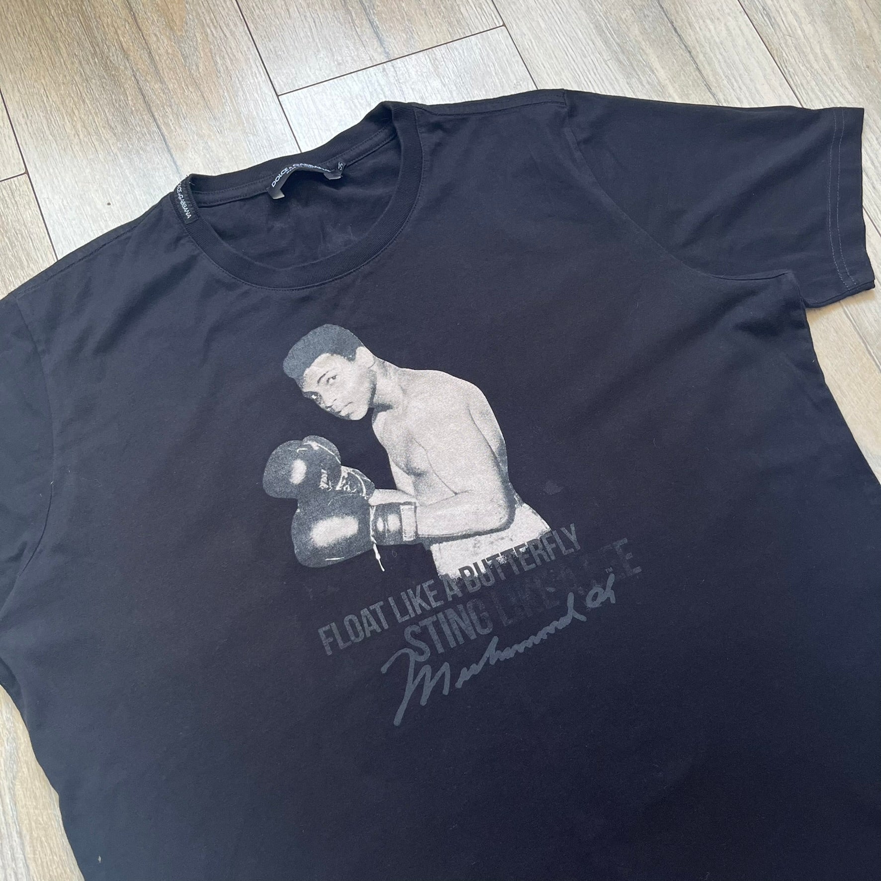  Dolce &amp; Gabbana Muhammad Ali Black Tee is a black T-shirt made in Italy from 100% cotton. The front features a grayscale portrait of Muhammad Ali. Beneath the image is the quote “float like a butterfly sting like a bee” along with a Muhammad Ali signature. 