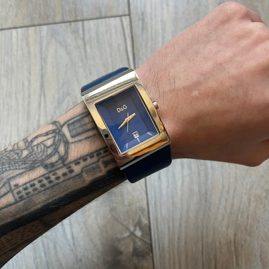 Dolce & Gabbana Navy & Silver Analog Watch features a silver-tone case with a navy dial and matching navy leather strap. The dial has a minimal design with silver hands, a small D&G logo, and a day-date window. The side of the case includes a raised Dolce & Gabbana logo, while the inside of the leather strap is stamped with D&G branding.