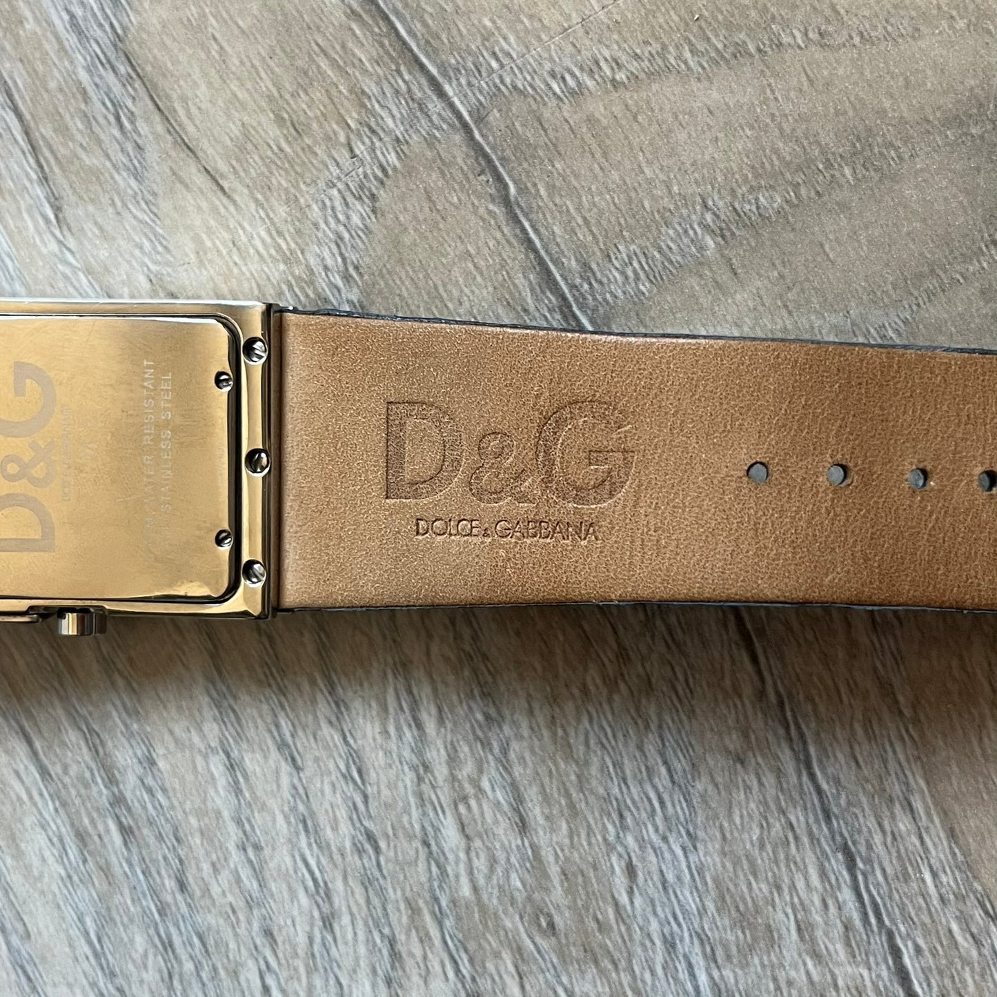 Dolce & Gabbana Navy & Silver Analog Watch features a silver-tone case with a navy dial and matching navy leather strap. The dial has a minimal design with silver hands, a small D&G logo, and a day-date window. The side of the case includes a raised Dolce & Gabbana logo, while the inside of the leather strap is stamped with D&G branding.