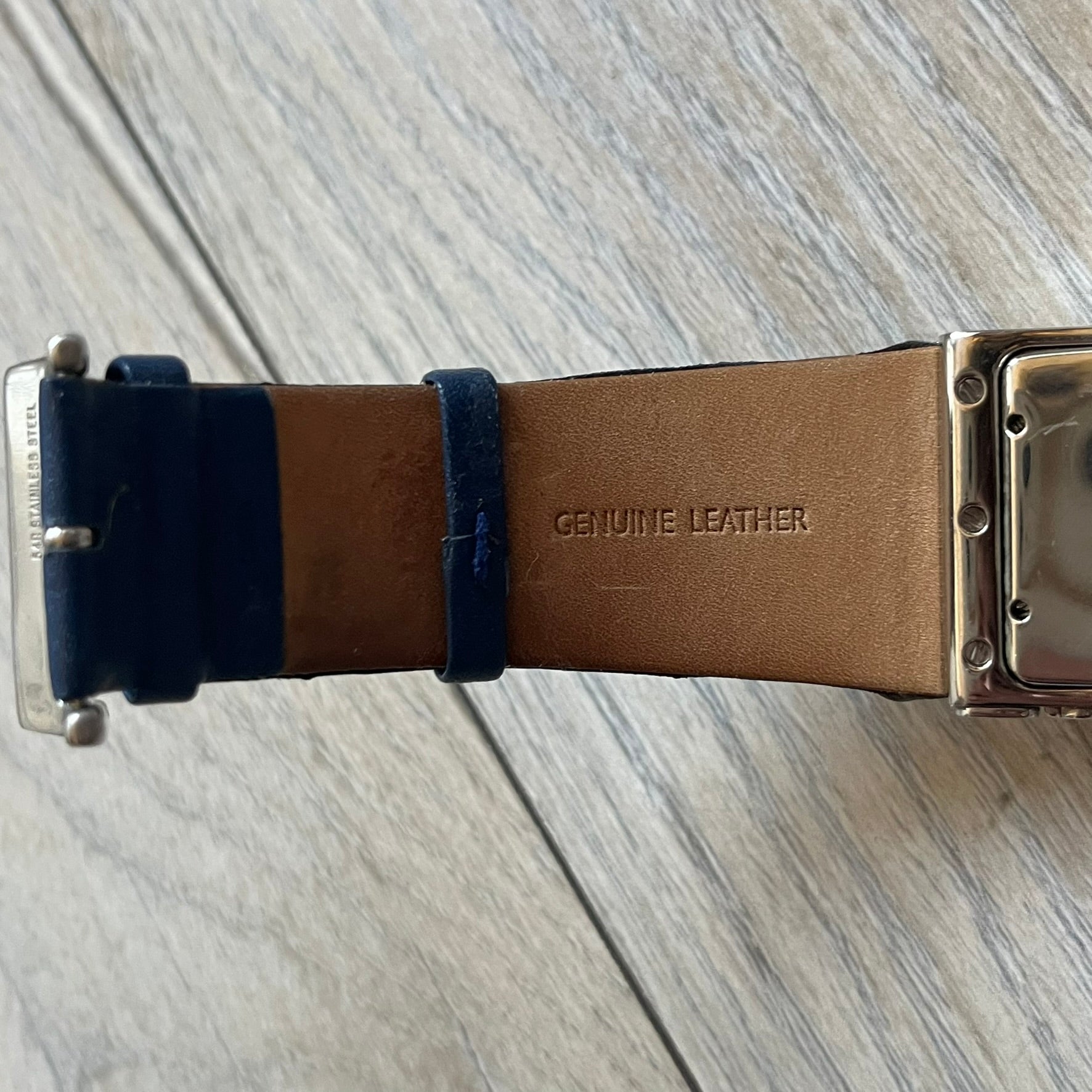 Dolce & Gabbana Navy & Silver Analog Watch features a silver-tone case with a navy dial and matching navy leather strap. The dial has a minimal design with silver hands, a small D&G logo, and a day-date window. The side of the case includes a raised Dolce & Gabbana logo, while the inside of the leather strap is stamped with D&G branding.