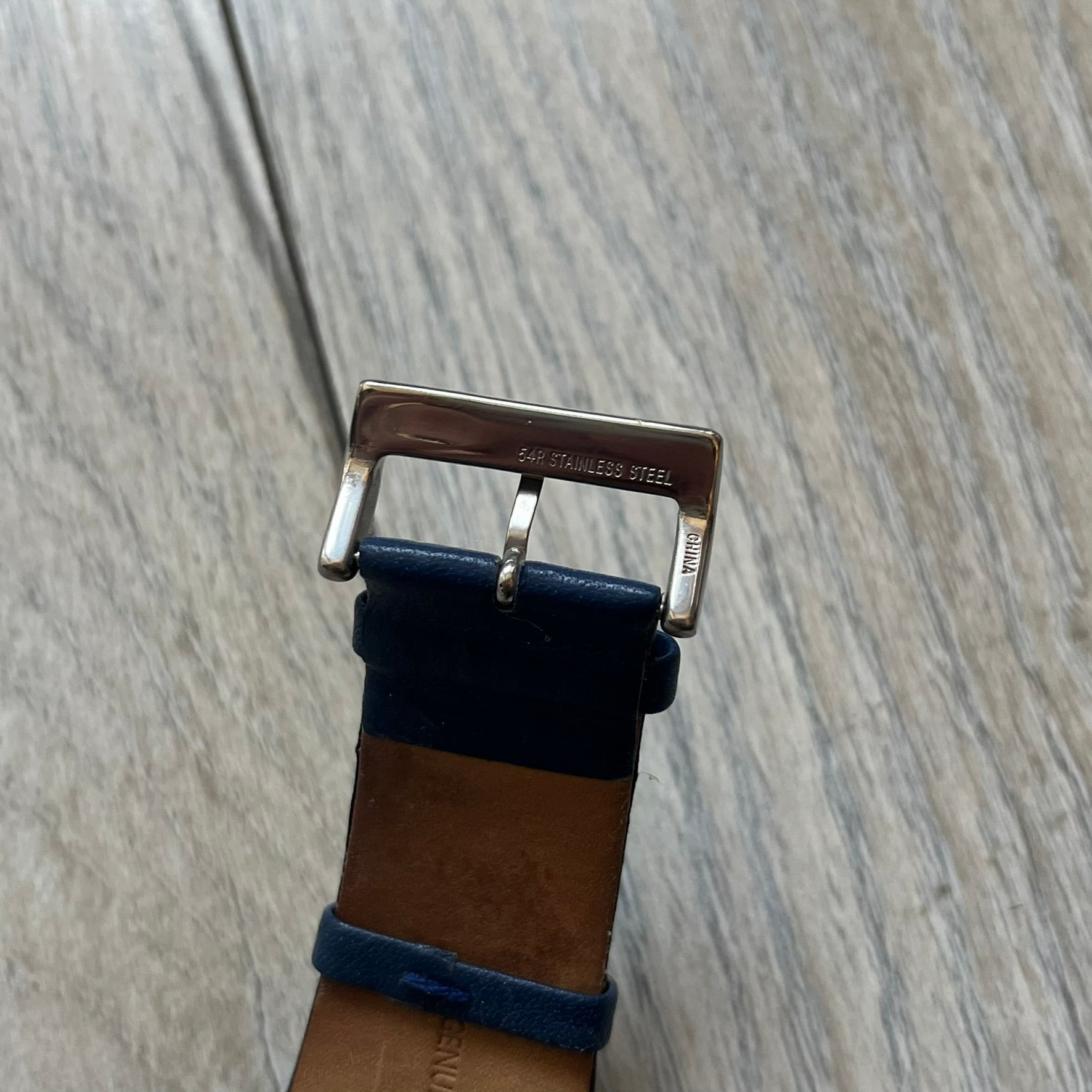 Dolce & Gabbana Navy & Silver Analog Watch features a silver-tone case with a navy dial and matching navy leather strap. The dial has a minimal design with silver hands, a small D&G logo, and a day-date window. The side of the case includes a raised Dolce & Gabbana logo, while the inside of the leather strap is stamped with D&G branding.